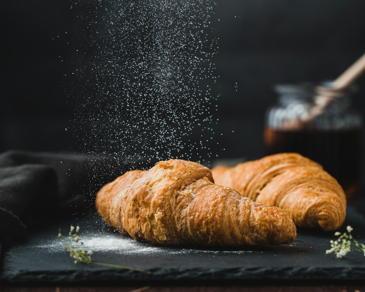 Two golden croissants dusted with flour on a dark surface